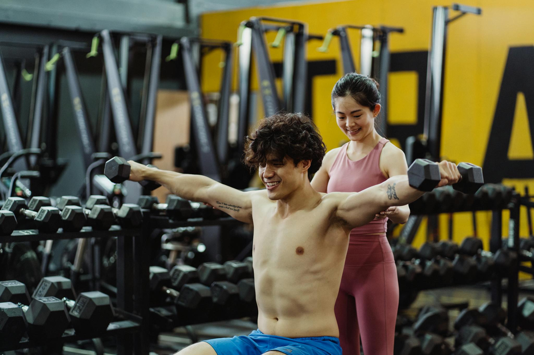 Man lifting weights with guidance from woman in modern gym setting.